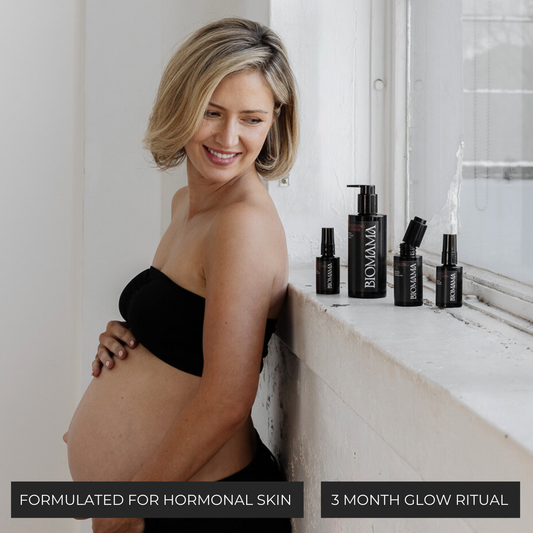 Pregnant woman smiling and holding her belly, with Biomama Essentials Set bottles on windowsill – luxury skincare formulated for hormonal skin.